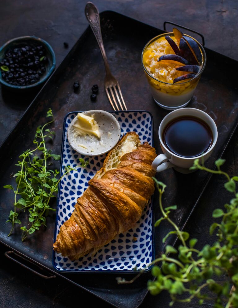 A cozy breakfast setup featuring a croissant, butter, coffee, and fruit yogurt on a tray.