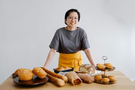 Smiling Asian female baker showcasing a variety of fresh bread and pastries indoors.